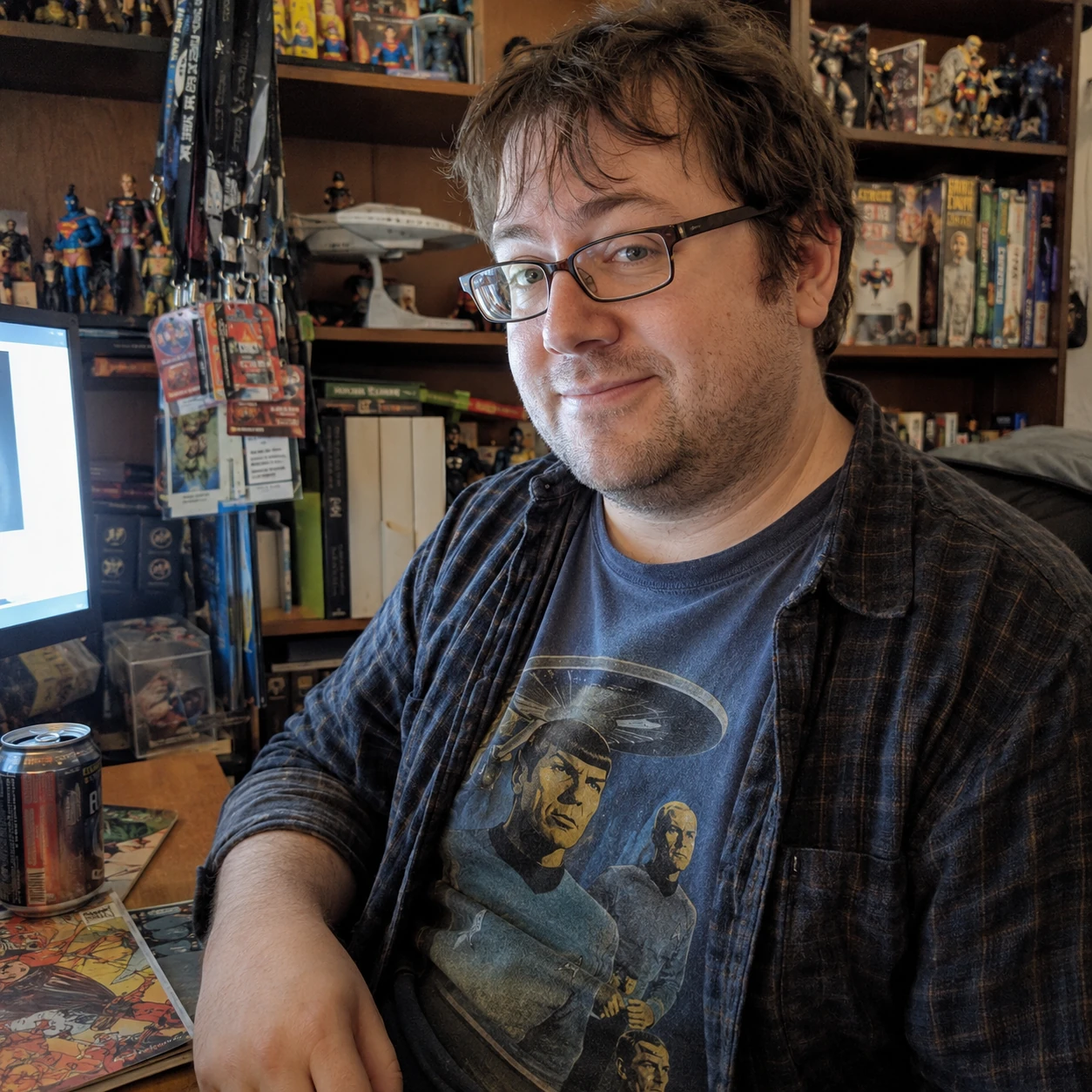 35-year-old man with glasses and a Star Trek shirt sitting at a cluttered computer desk surrounded by comics and collectibles.